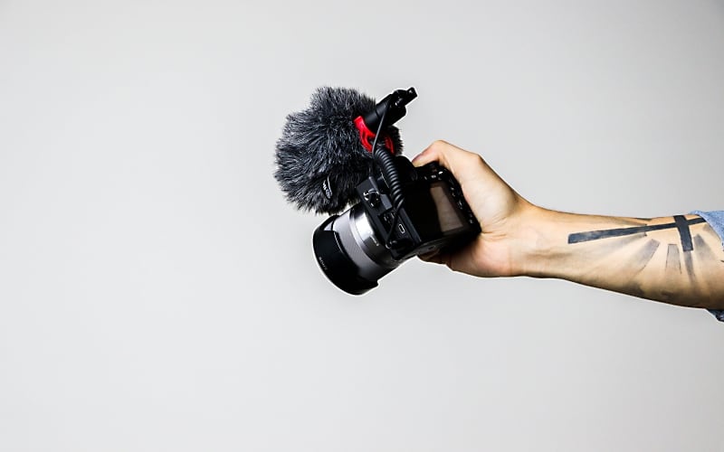 An ausgestrecktem Arm gehaltene Siegelreflexkamera mit aufgesetztem Mikrofon samt Windschutz