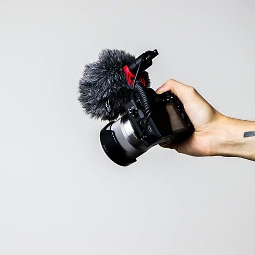 An ausgestrecktem Arm gehaltene Siegelreflexkamera mit aufgesetztem Mikrofon samt Windschutz